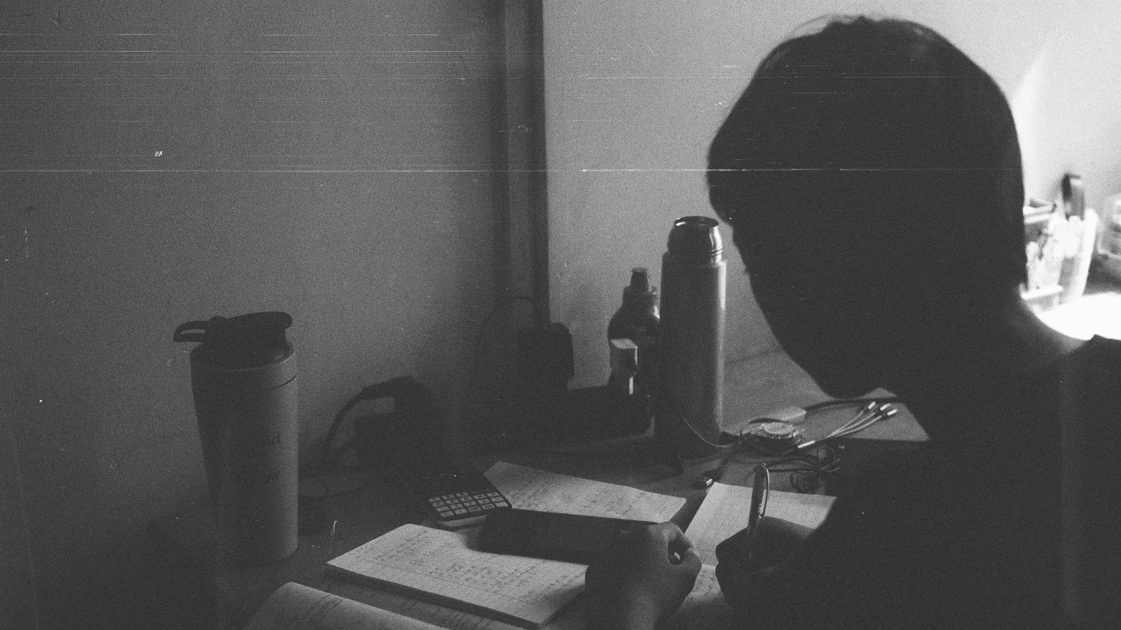 어두운 책상에서 종이에 글을 쓰고 있는 사람, 커피잔과 물병이 보임