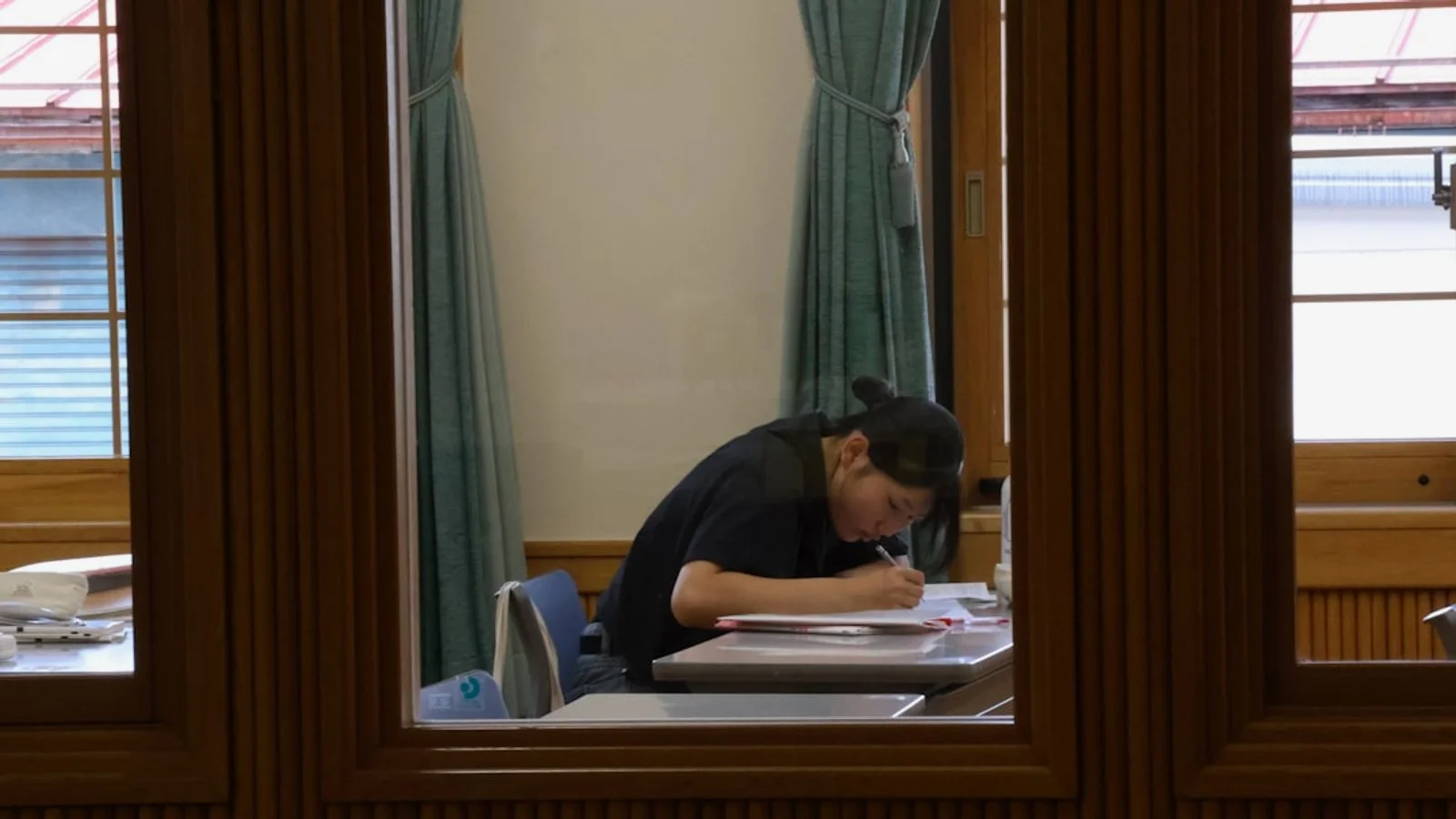 창문이 있는 실내에서 검은색 옷을 입은 사람이 책상에 앉아 서류에 글을 쓰고 있다.